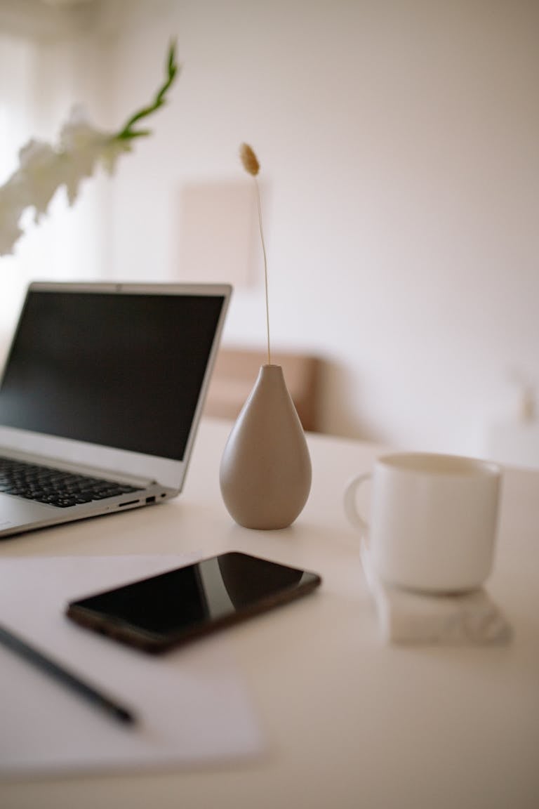 Minimalist workspace with a laptop, coffee mug, and a simple vase on a white desk, reflecting calm, organized bookkeeping support for small businesses.