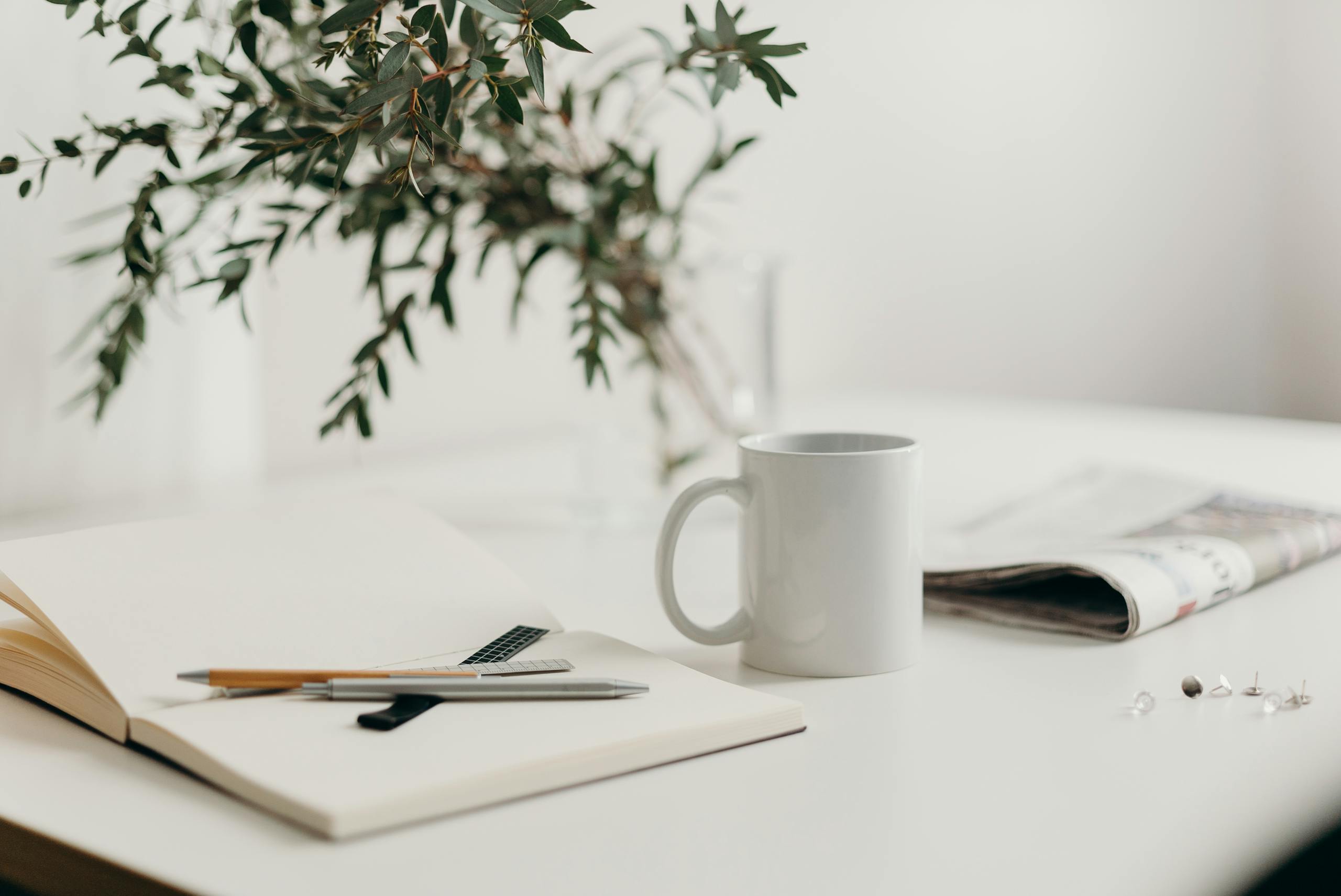 A calm bookkeeping workspace with a notebook, pen, coffee mug, and greenery on a white desk