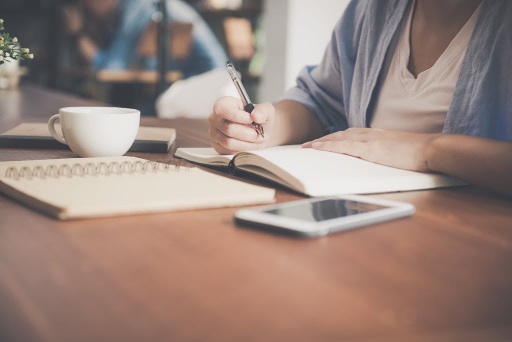 Person writing in a notebook at a wooden desk with a smartphone and coffee—focused, modern bookkeeping workspace
