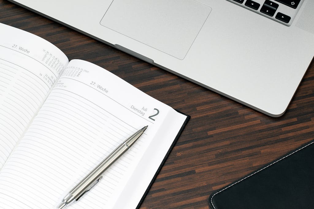 A home office setup for monthly bookkeeping services featuring a laptop with a digital calendar, a blank notebook, and a cup of black coffee on a beige throw—ideal environment for organized small business bookkeeping and QuickBooks Online management.