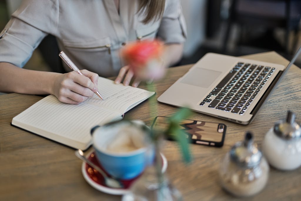 Woman taking notes beside a laptop and coffee in a cozy workspace, symbolizing judgment-free QuickBooks Online coaching and remote learning for small business owners.